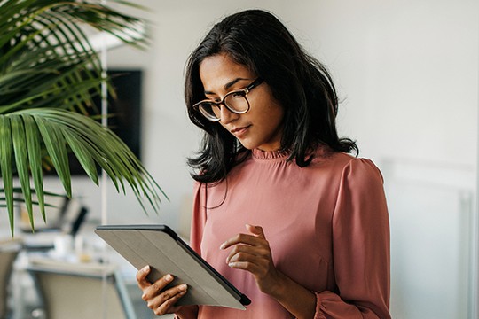 A woman with glasses is focused on a tablet, tapping the screen with one hand. She is surrounded by indoor greenery and a modern office environment in the background.