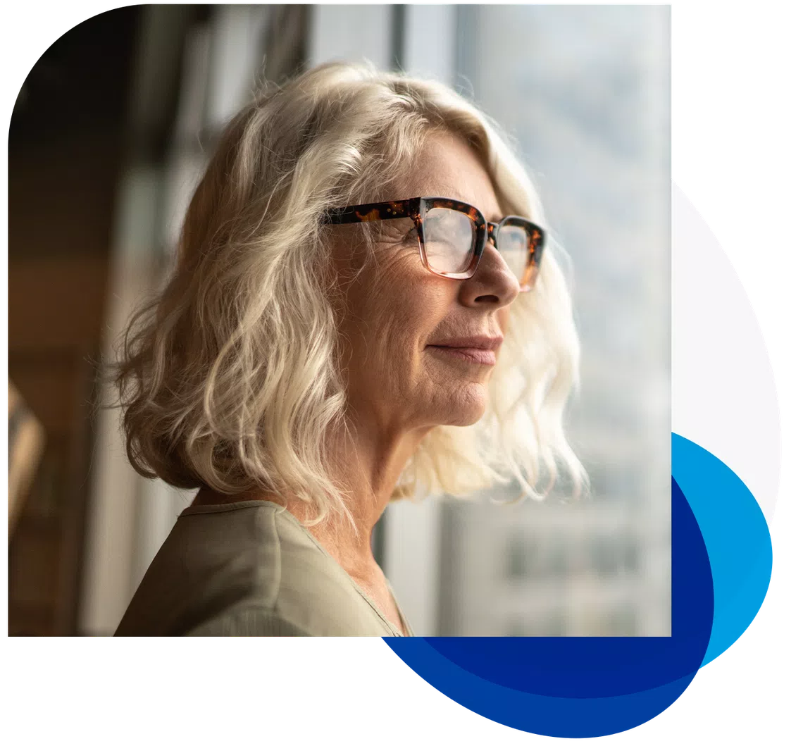 A woman with curly, blonde hair and glasses gazes thoughtfully out a window, bathed in soft natural light, with blurred interior surroundings suggesting a modern space.
