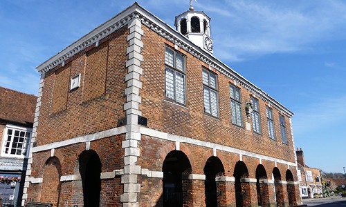 A brick building with a clock tower stands prominently. It features large windows and supportive arches beneath. The setting includes a clear blue sky, suggesting a serene outdoor environment.