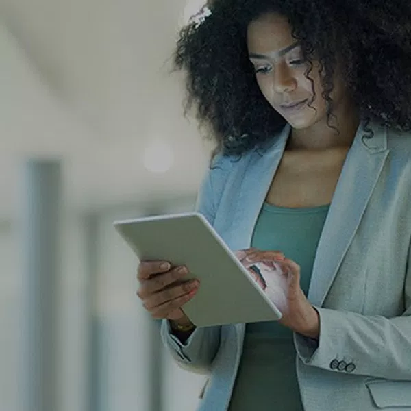 A woman with curly hair, dressed in a blazer, is engaged with a tablet. She stands in a modern interior space, with blurred columns and soft lighting in the background.