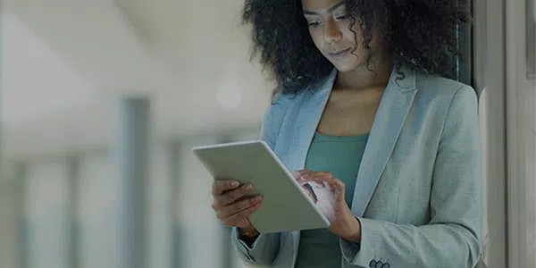 A woman with curly hair, dressed in a blazer, is engaged with a tablet. She stands in a modern interior space, with blurred columns and soft lighting in the background.