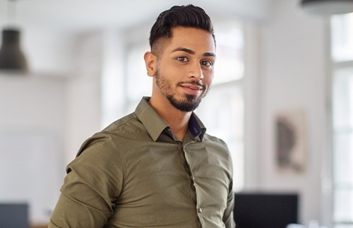 A man stands confidently in a bright office space, wearing a green shirt. He has short dark hair and a slight smile, surrounded by blurred workstations and natural light.