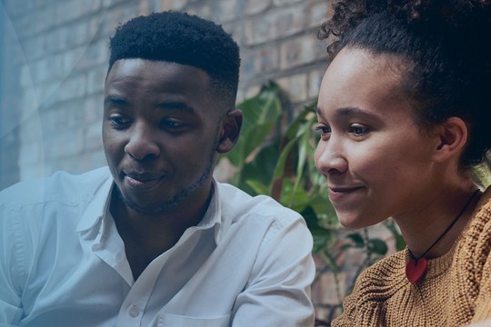 A man and a woman are sitting together, focused on a digital tablet. They appear engaged and are surrounded by greenery, indicating a relaxed indoor setting.