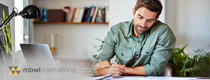 A man wearing a green shirt writes thoughtfully at a desk, surrounded by plants and bookshelves in a bright, inviting workspace. The scene conveys focus and creativity.