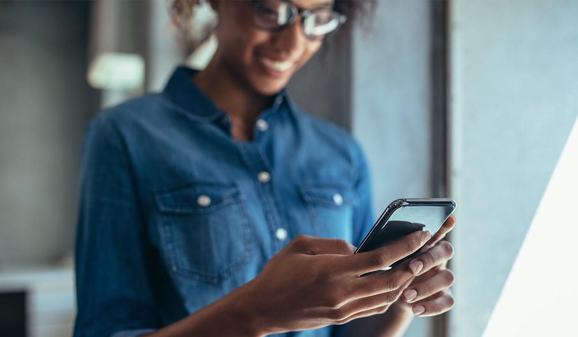 A woman in a denim shirt smiles while looking at her smartphone. She stands indoors, with soft natural light illuminating the space around her, creating a relaxed atmosphere.
