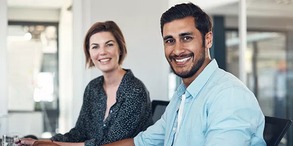 Two smiling individuals sit at a modern workspace, engaged in a friendly conversation. The context features an open office environment with natural light coming through large windows.
