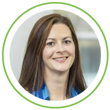 A woman with long, straight brown hair smiles while wearing a blue shirt. The background is blurred, featuring soft colors that suggest an indoor environment.