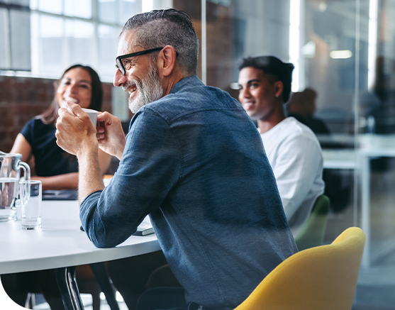 A middle-aged man with glasses smiles while holding a coffee cup at a round table. Two other people engage in conversation, surrounded by a bright, modern office environment.