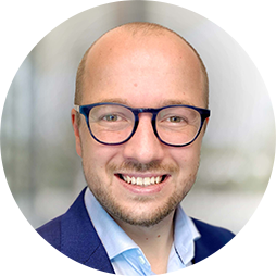 A man with short hair and glasses smiles confidently, wearing a dark blazer over a light shirt. The background is softly blurred, suggesting a professional environment.