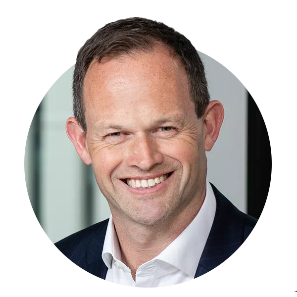 Smiling man wearing a dark suit and white shirt stands in a modern office setting with glass and neutral tones in the background, conveying a professional and approachable demeanor.