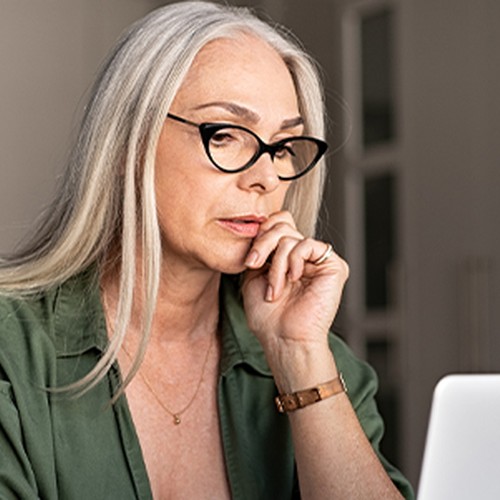 A woman with long gray hair and glasses is thoughtfully gazing at a laptop, seated at a modern table in a bright, minimalistic room with plants and soft furnishings.