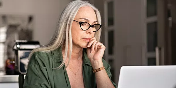 A woman with long gray hair and glasses is thoughtfully gazing at a laptop, seated at a modern table in a bright, minimalistic room with plants and soft furnishings.