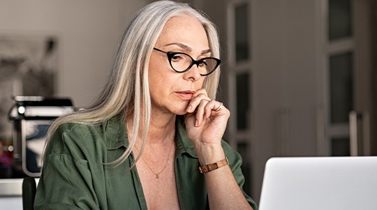 A woman with long gray hair and glasses is thoughtfully gazing at a laptop, seated at a modern table in a bright, minimalistic room with plants and soft furnishings.