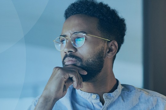A man with glasses is thoughtfully resting his chin on his hand, gazing out a window. The background features soft blue tones, creating a calm and contemplative atmosphere.