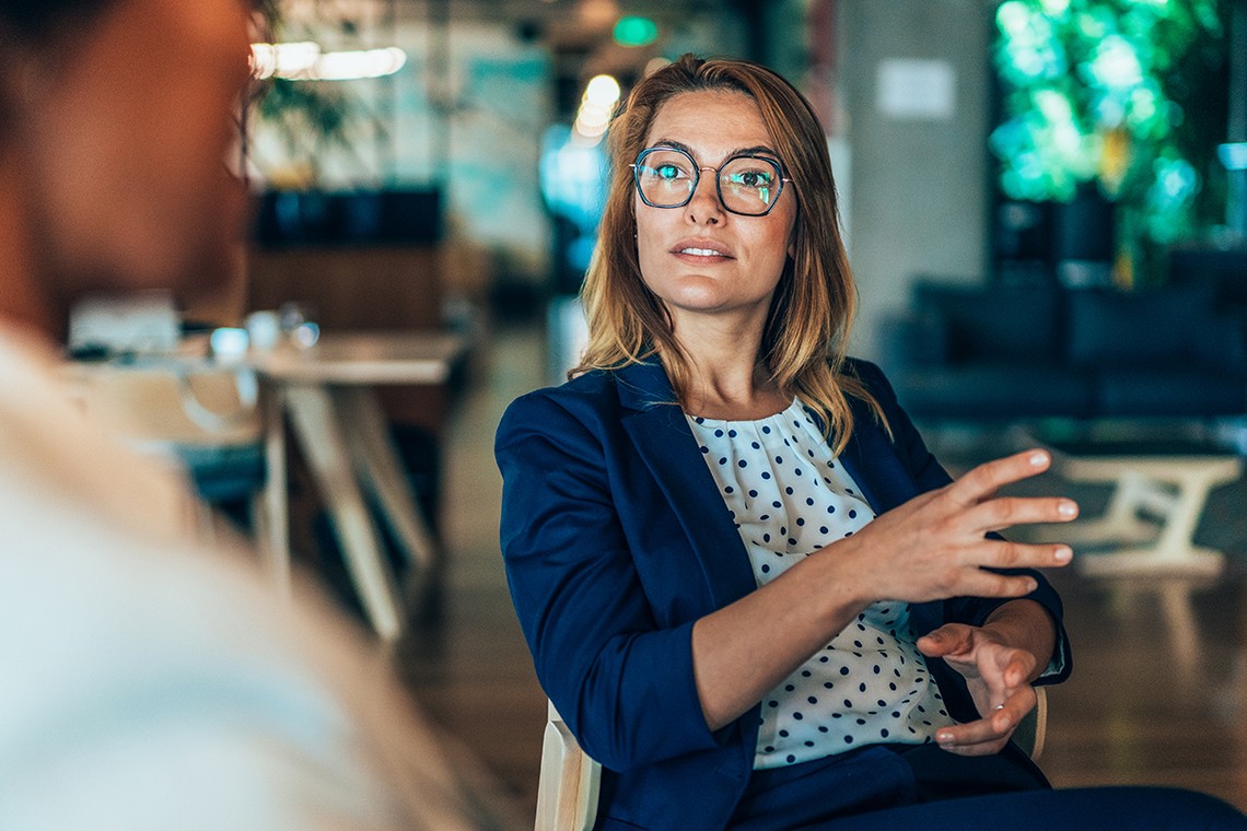 A woman in a blue suit with glasses gestures while speaking in a modern, well-lit office environment, sitting across from another person who is out of focus.