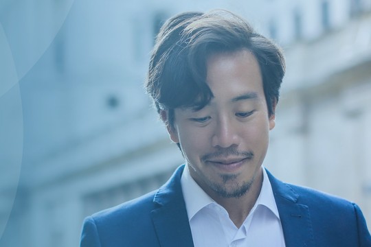 A man in a blue suit is looking down, appearing contemplative. The background features blurred architectural elements, suggesting an urban setting with soft, muted colors.
