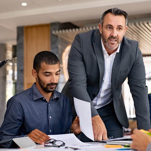 A man in a suit stands by a table, gesturing while discussing documents with three seated colleagues, in a modern office environment filled with natural light.