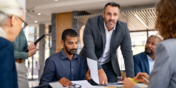 A man in a suit stands by a table, gesturing while discussing documents with three seated colleagues, in a modern office environment filled with natural light.