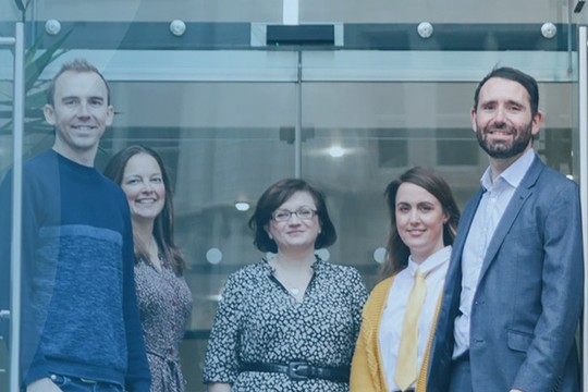Five individuals stand in front of a glass entrance, smiling and posing together. The setting features modern architecture and greenery, creating a welcoming atmosphere.
