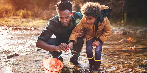 A man and a child crouch in shallow water, using a net to catch small creatures. The sun sets in the background, reflecting warm colors on the water's surface.