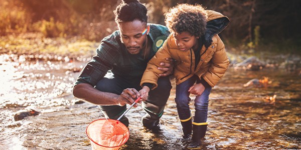 A man and a child crouch in shallow water, using a net to catch small creatures. The sun sets in the background, reflecting warm colors on the water's surface.