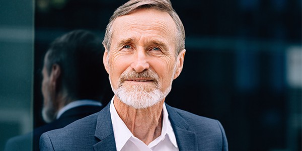 A mature man with a gray beard and hair wears a dark suit over a white shirt, smiling. He stands in front of a reflective surface, suggesting an urban environment.