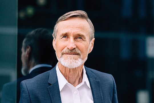 A mature man with a gray beard and hair wears a dark suit over a white shirt, smiling. He stands in front of a reflective surface, suggesting an urban environment.