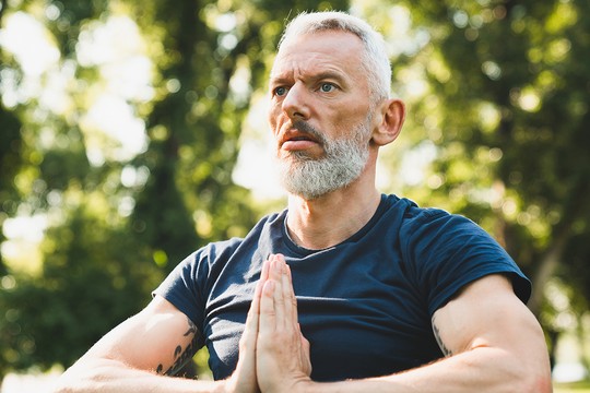 A man with a beard and short gray hair is practicing meditation outdoors, sitting cross-legged with hands in a prayer position, surrounded by green trees and soft sunlight.