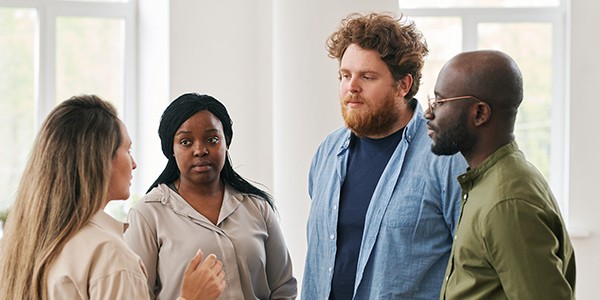 A group of four individuals stands in a bright, open space, engaging in conversation. One person speaks animatedly, while the others listen attentively, displaying various expressions.
