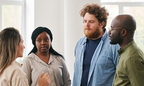 A group of four individuals stands in a bright, open space, engaging in conversation. One person speaks animatedly, while the others listen attentively, displaying various expressions.