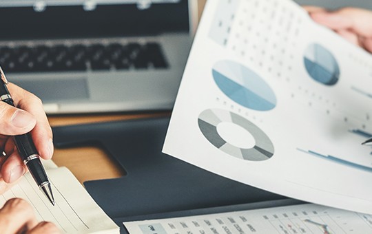 Two hands hold documents featuring graphs and charts, while a laptop is in the background. The setting suggests a collaborative environment focused on data analysis and discussion.