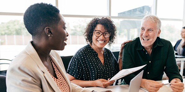 Three individuals are engaged in a lively conversation at a table. One person is presenting a document while others are smiling and actively listening in a bright, modern office environment.