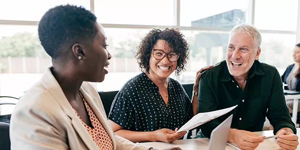 Three individuals are engaged in a lively conversation at a table. One person is presenting a document while others are smiling and actively listening in a bright, modern office environment.