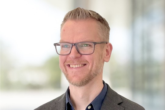 A man with short, light brown hair and glasses smiles while wearing a dark blazer over a collared shirt. The background features soft, blurred greenery, suggesting an outdoor or open space.