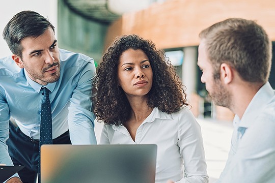 A laptop is placed on a table as three professionals discuss, with a focus on a woman listening attentively. The setting is a modern, bright office space.