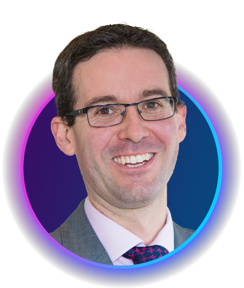 A man with glasses smiles broadly, wearing a gray suit and a patterned tie. His portrait is displayed within a circular design featuring gradient colors of purple and blue.