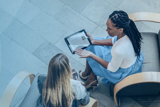 Two women are seated in a modern, spacious environment. One woman is using a tablet, while the other is engaged in discussion. The floor is light-colored and features a plant nearby.