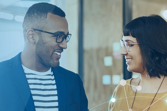 Two people, a man and a woman, are smiling and conversing in a modern indoor setting. The background features soft colors and abstract shapes, suggesting a friendly atmosphere.