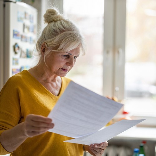 A woman in a yellow sweater examines several sheets of paper in a bright kitchen, surrounded by a refrigerator adorned with magnets and a sunlit window showing an outdoor scene.
