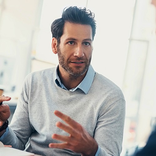 A man in a light sweater gestures animatedly while speaking, holding a pen, as he engages with another person in a bright, modern office setting with large windows.