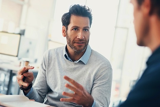 A man in a light sweater gestures animatedly while speaking, holding a pen, as he engages with another person in a bright, modern office setting with large windows.