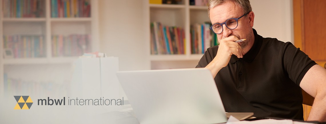 A man in a black polo shirt is focused on a laptop, thoughtfully holding a pen to his mouth. Behind him are shelves filled with colorful books, indicating a home office environment.