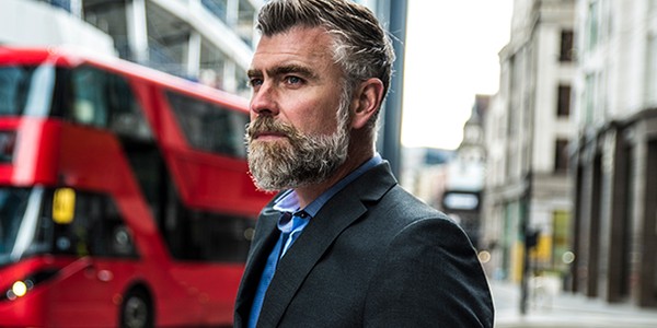 A well-dressed man with a beard stands confidently on a city street, looking thoughtfully towards the side. A red double-decker bus passes behind him in an urban setting.