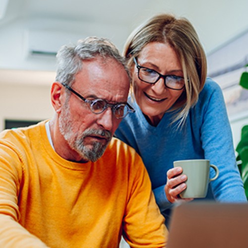 An older man in an orange sweater and glasses is intently looking at a laptop, while a woman in a blue sweater holds a mug and smiles, both sitting in a cozy, well-lit room.