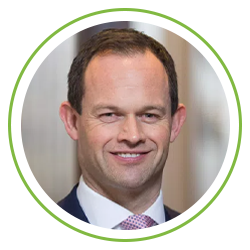A man with short hair and a slight smile is dressed in a suit and tie, set against a blurred interior background, suggesting a professional or corporate environment.