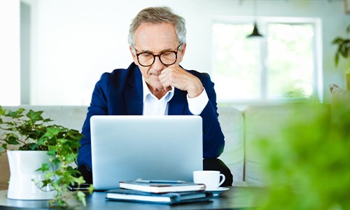 A senior man sits thoughtfully in front of a laptop, resting his chin on his hand. Surrounded by greenery, he is in a bright, modern indoor space, with books and a coffee cup on the table.