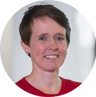 A person with short dark hair smiles while wearing a red shirt, standing in a bright, modern environment with soft-focus background elements suggesting an office setting.
