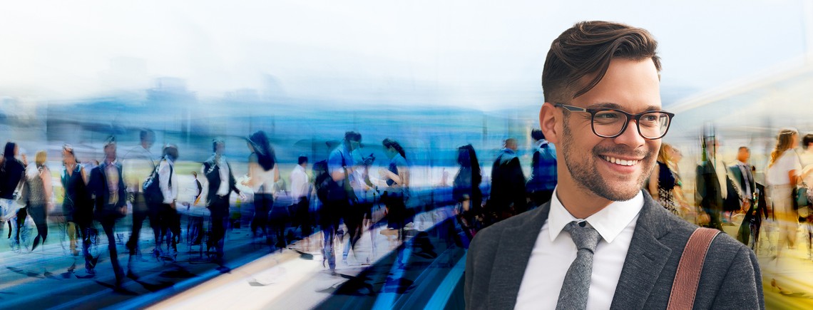 A smiling man in a suit, wearing glasses, stands in focus with a blurred crowd of people walking past in a busy urban environment.