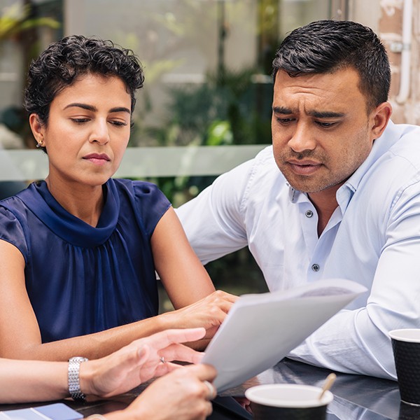 A woman and a man sit closely at a table, focused on a document. Their expressions are serious as they discuss its contents in a well-lit outdoor setting with greenery in the background.