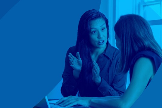 Two women are engaged in a conversation, with one actively gesturing while speaking. They are seated at a table, surrounded by a bright, blue-toned background.